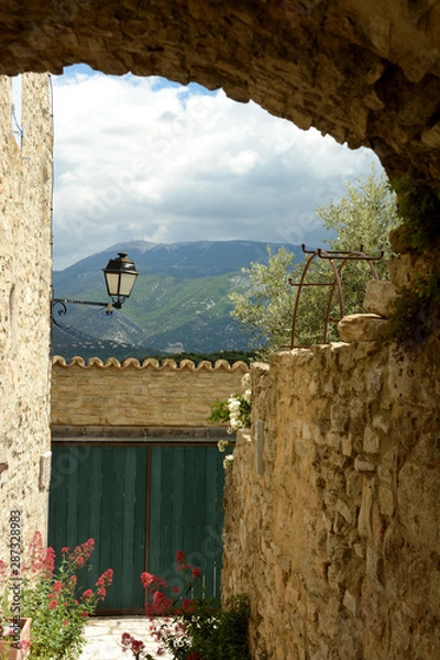 Obraz View of the Mont Ventoux through a porch of the village of Faucon. Ventoux mountain. In the foreground a street light, a door, flowers, a tree, a facade. Spring in France, Provence, Vaucluse.