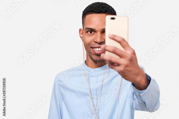 Fototapeta Image of happy African Amerfican young man holds modern smart phone, speaking online and blogging on cellphone. Handsome Afro male smiling and making self portrait on his device. People and technology