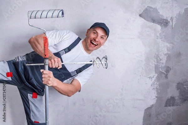 Fototapeta Uśmiechnięty pracownik w kombinezonie i czapce trzymający w rękach dyszę do miksera elektrycznego pokrytego kitem i wałek do malowania. Naprawa koncepcji pracy, budowy i zawodu. Skopiuj miejsce