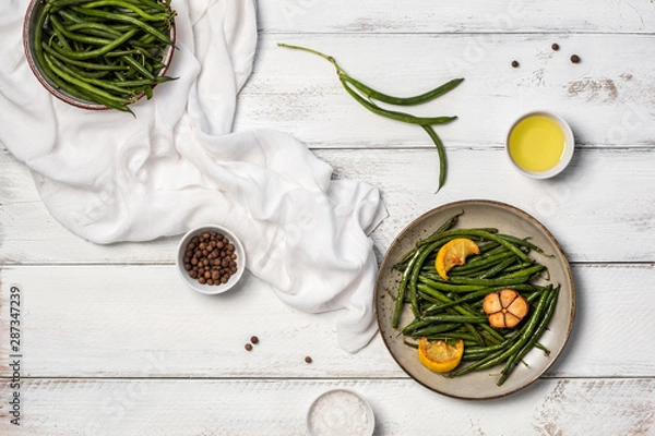 Fototapeta Plate with fried green beans, lemon pieces and garlic, uncooked pods, small bowls with olive oil, sea salt and peppercorns and towel on white wooden table. Healthy food concept. Flat lay, top view