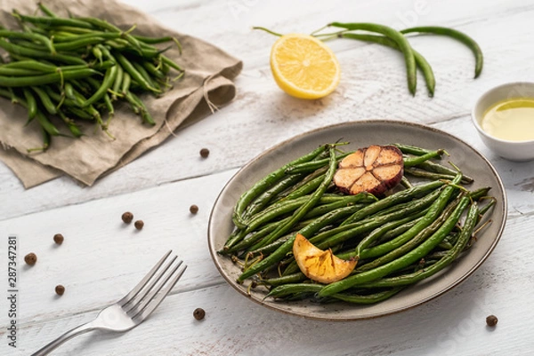 Fototapeta Plate with fried green beans, lemon piece and garlic, uncooked pods on linen napkin, small bowl with olive oil, peppercorns, lemon and a fork on white wooden table. Healthy food concept