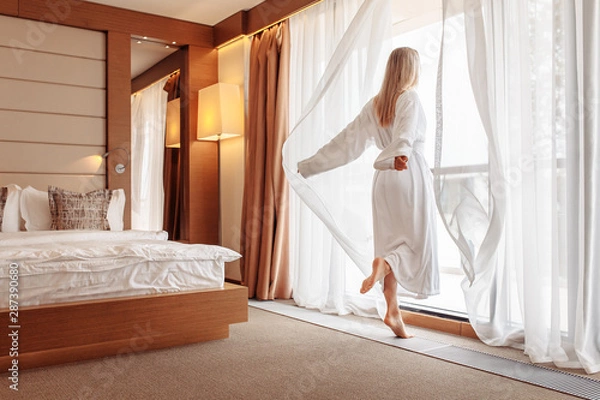 Obraz Joyful emotional young beautiful woman in white bathrobe happily opens the curtains of her window in the bedroom on a sunny summer morning. The concept of starting a new day and fulfilling your plans