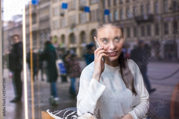 Obraz View through coffee shop window with street reflection of a hipster girl having mobile phone conversation with friend while sitting in restaurant. Woman talking on cell telephone during rest in cafe