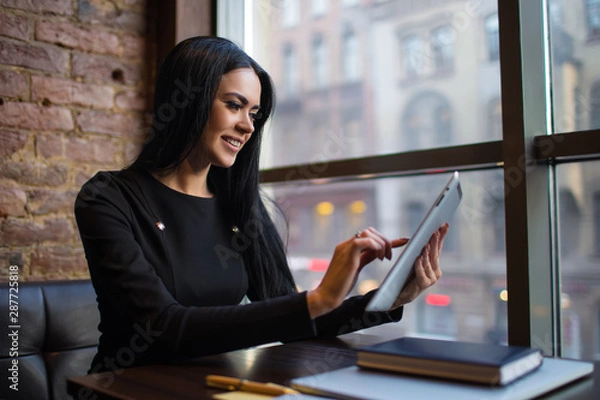 Obraz Confident woman intelligent government worker checking e-mail on portable touch pad computer while sitting at workplace in office interior. Female economist using digital tablet for online chat  