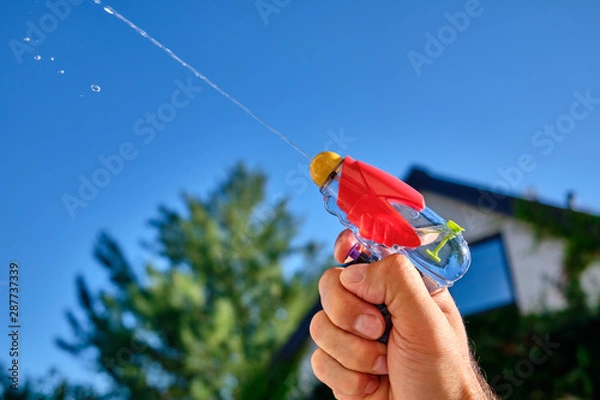 Obraz Colorful water pistol spraying out a jet of water in a caucasian male hand in front of  a house and the blue sky of a sunny summer day