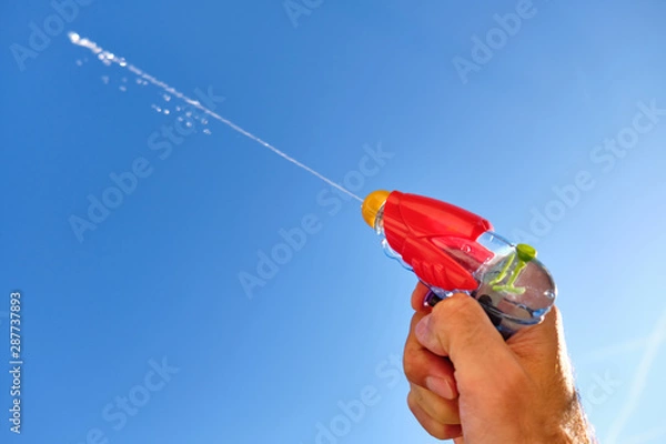 Obraz Colorful water pistol spraying out a jet of water in a caucasian male hand in front of  the blue sky of a sunny summer day