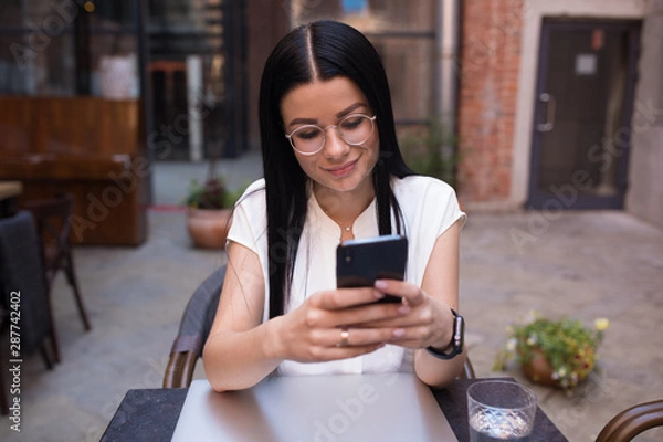 Fototapeta Fashionable female skilled lifestyle blogger reading article on web site via mobile phone while sitting in coffee shop during recreation time. Woman student typing text on cell telephone