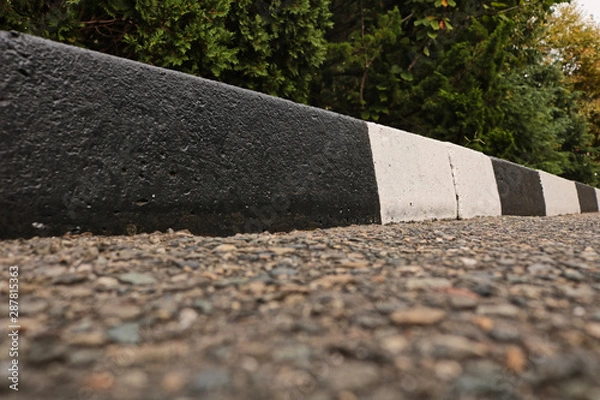 Fototapeta A street border painted in white and black on the background of the road and green foliage in the summer closeup. Road safety