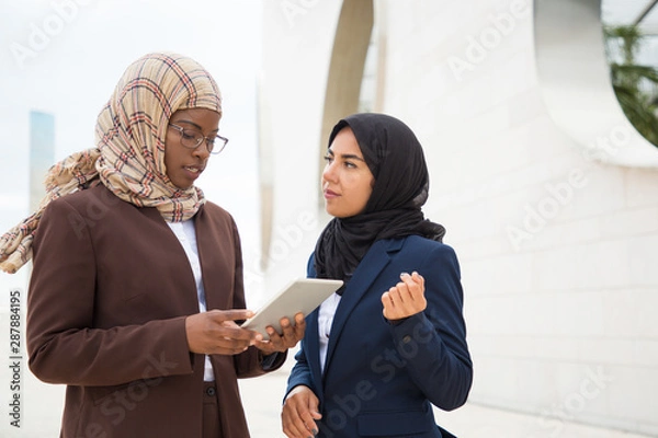 Fototapeta Focused Muslim business woman explaining project specifics to colleague. Business women in hijabs standing outdoors, holding tablet and talking. Expertise concept