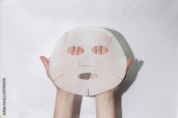 Fototapeta  Sheet face mask. Fabric mask for facial skin. Female hands hold a face mask. On a white background. View from above. Cosmetology, medicine and healthcare. The concept of home treatments for moisturiz