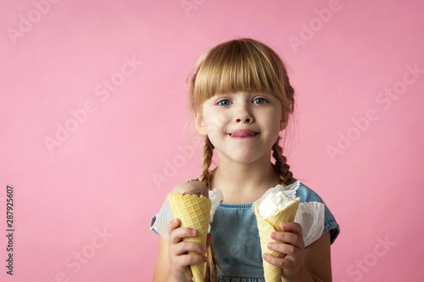 Fototapeta little girl with pigtails in a blue dress eating ice cream in a cone on a pink background