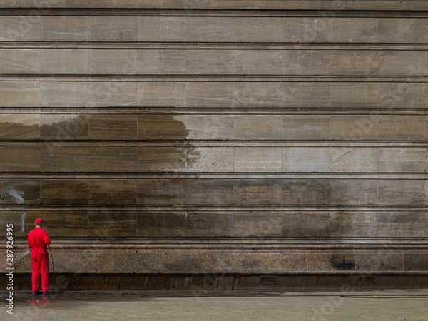 Obraz Unidentified man in red clothes washing graffiti from a building wall with high pressure water machine