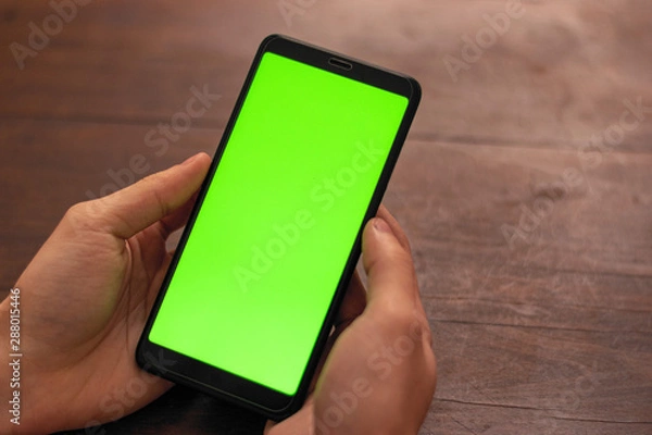 Fototapeta Hand of caucasian person holding a black unidentified smartphone with green screen on a wooden table background