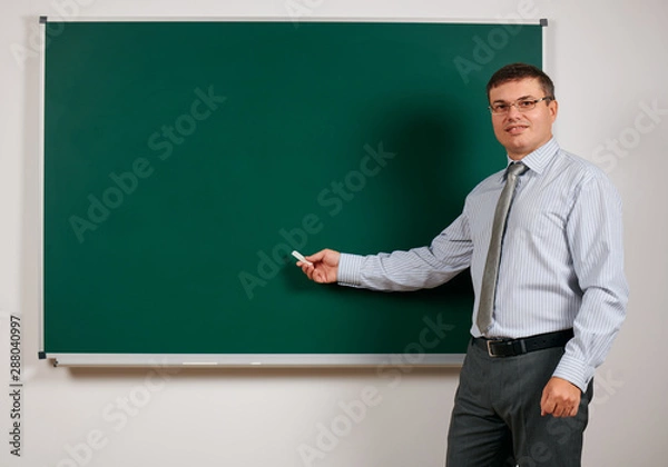 Fototapeta Portrait of a man dressed as a school teacher in business suit, posing at blackboard background - learning and education concept