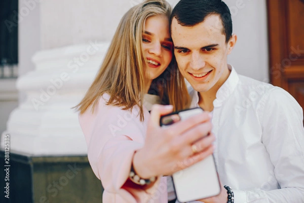 Fototapeta beautiful and stylish blonde along with her handsome guy standing near building in the summer city and use the phone in white case