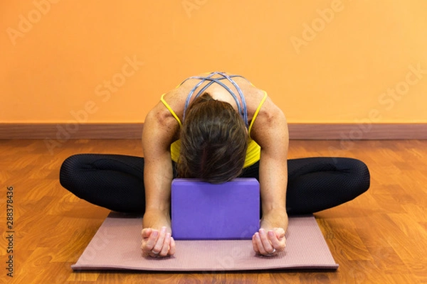 Fototapeta Woman in butterfly yin yoga asana with forehead resting on purple prop. Female yogi on baddha konasana pose on wooden plank floor studio. Relax exercise, flexibility concepts