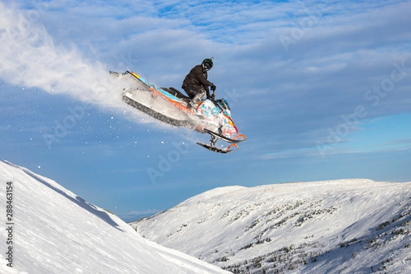 Obraz a snowmobile flies in the mountains with a large snow trail against a bright blue sky with clouds. Snowmobile jump at high altitude. bright snowmobile and looking at the camera. super quality photo