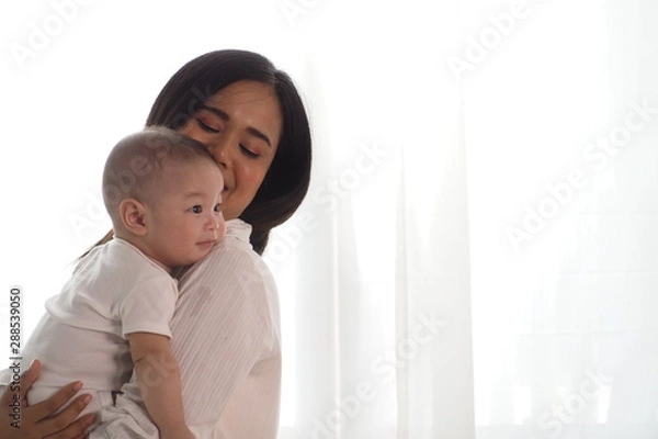 Fototapeta Beautiful Asian mother cradling her newborn baby on shoulder with happy face of being motherhood at home with white curtains background. Warm moment of mother and child, love and lifestyle concept