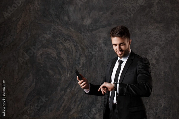 Fototapeta a young bearded man looks at his watch and holds a mobile phone in his hands. successful businessman in a suit watches time on a wristwatch