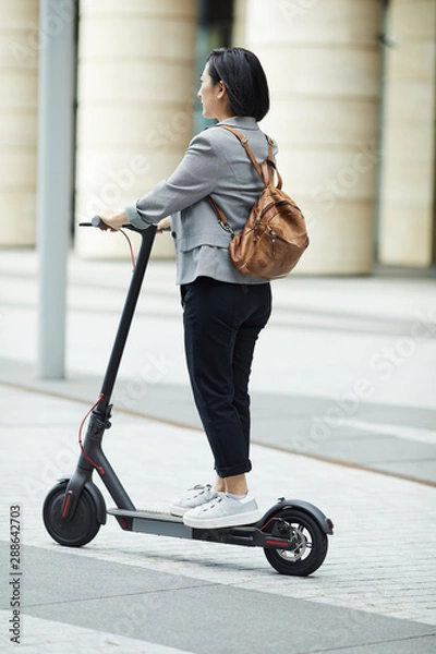 Fototapeta Full length side view of young Asian woman riding electric scooter in city street with architecture in background, copy space