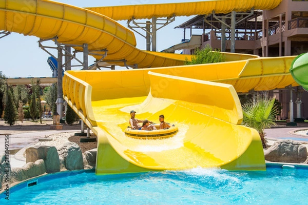 Fototapeta On a bright sunny day at the water park, the family slides down the hill on an air mattress.