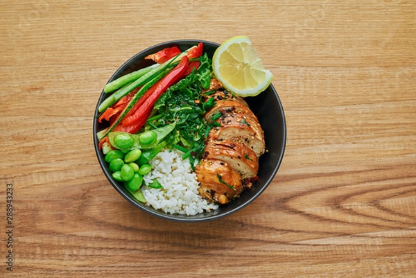 Obraz Teriyaki chicken fillet, fresh cucumber, red bell pepper, stew cabbage, edamame and basmati rice in black bowl. Traditional Asian Buddha bowl meal. Top view, directly above shot.