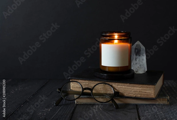 Obraz candle burning on stacked books, reading glasses, crystal, antique books, burning scented candle with blank label, copyspace, dark background with copy space, autumn mood, candle label design mock up