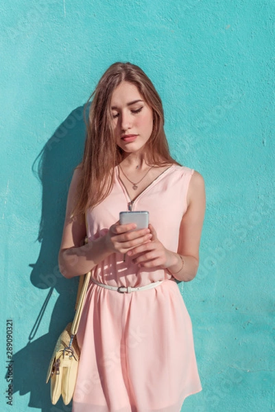 Fototapeta Beautiful girl in pink dress, background turquoise blue wall, in hands of mobile phone, reads and writes a message, online application on wifi social networks. Yellow bag. In summer in city.