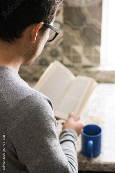 Obraz An unrecognizable young man reads a book and drinks a cup of coffee at home