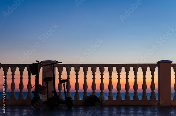 Fototapeta Two with a backpack and a women's bag tourists left near the fence with balusters against the sunset by the sea.