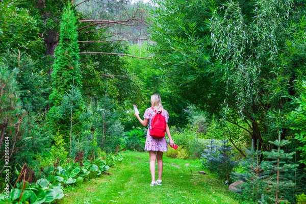Fototapeta A view from the back of a young blonde girl in a pink dress with a red backpack on her back, a phone in her hand walks along a path and looking to the map in a park or forest. Travelling in wild.