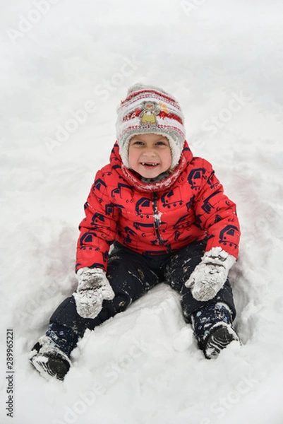 Obraz A child in a red jacket playing in the snow. The cute boy walks in the winter on the street. The kid laughs and looks at the camera. Happy childhood, fun weekend in nature. It snowed,winter is coming.
