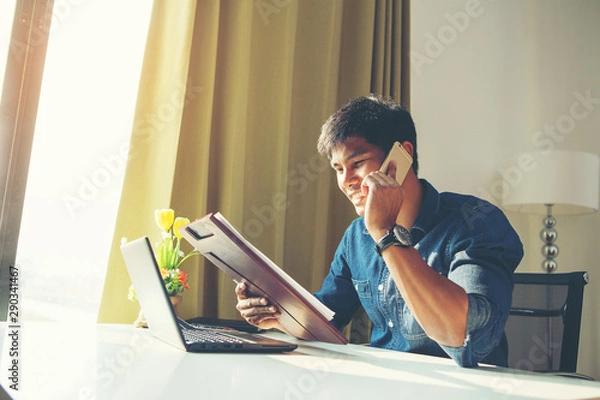 Fototapeta Attractive asian guy talking on phone while using laptop at workplace.