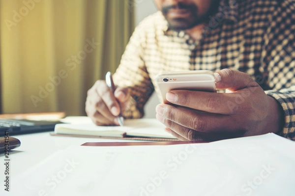 Fototapeta Casual business man using mobile smart phone working, taking note, schedule on paper notebook, reading e book application. E learning concept