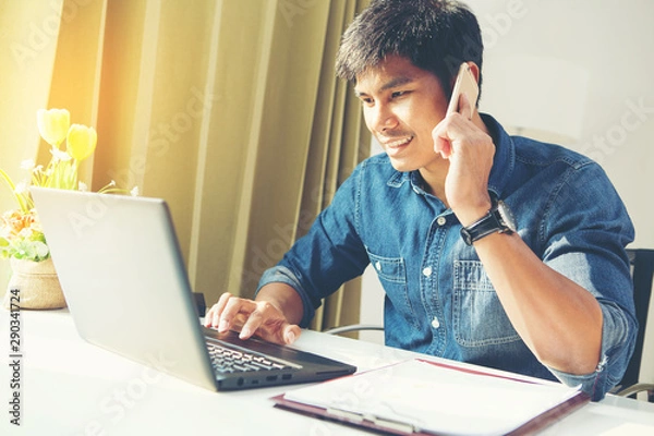 Fototapeta Attractive asian guy talking on phone while using laptop at workplace.