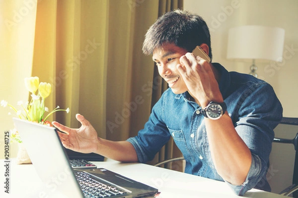 Fototapeta Attractive asian guy talking on phone while using laptop at workplace.