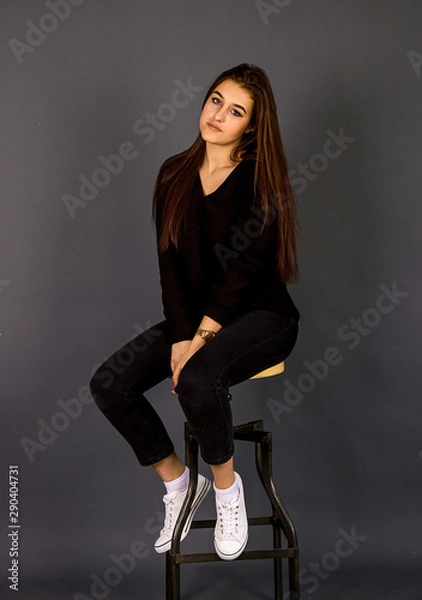 Fototapeta Young, beautiful girl in black clothes and white gym shoes.  A girl sits in a beautiful pose near a high chair, shooting in the studio on a plain background.
