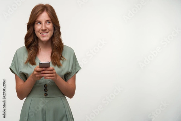 Fototapeta Lovely young redhead female with smartphone in hands plotting something good, looking aside and biting underlip, posing over white background
