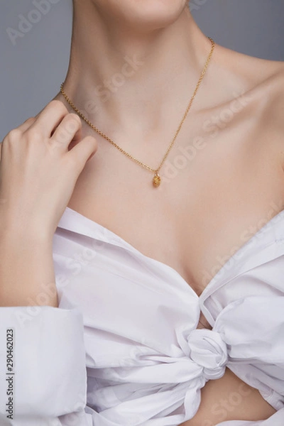 Fototapeta Cropped shot of a lady, wearing white chest tied shirt. She has golden chain with a pendant on her neck. Her right hand is touching shoulder. The photo is taken by grey background. 