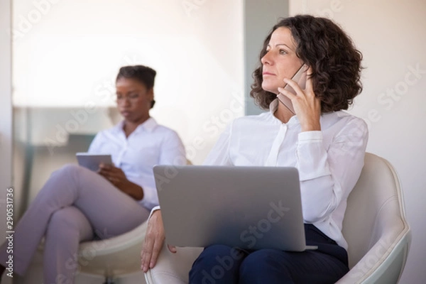 Fototapeta Serious excited businesswoman talking on phone at computer. Business woman sitting in armchair, using laptop and calling on cellphone in office lobby. Communication concept