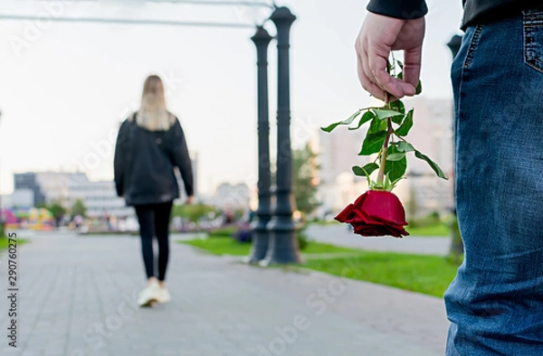 Fototapeta View of the red rose flower in the hand of the guy on the background of the girl leaving him in the city Park, square. Separation, resentment, quarrel, dislike, unloved