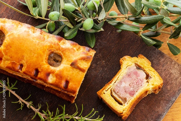 Fototapeta Top view of a pate en croute or pâté en croûte, with rosemary twig and green olives on branch with leaves over a dark wooden cutting board background. French traditional appetiser.