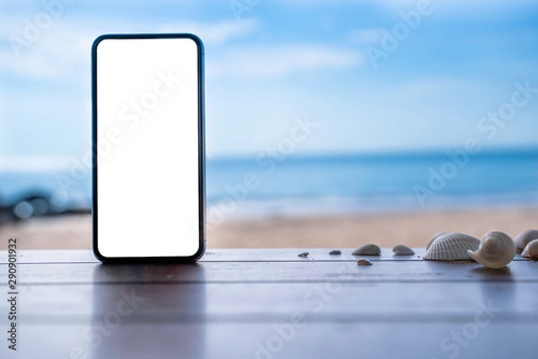 Fototapeta stock photo phone in the beach, black smartphone show white screen placed on wooden table with shells. Sky summer,beach and sea in background with copy space , blank for text ads and graphic design.
