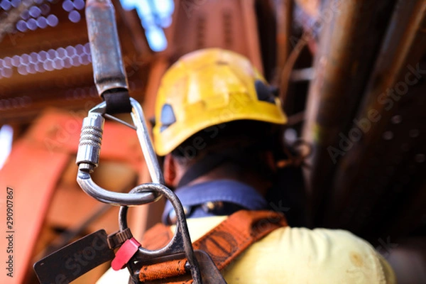 Obraz Construction worker welder wearing safety helmet, fall arrest harness clipping locking Karabiner attached retraceable energy shock absorber lanyard device on the back of his safety harness loop 