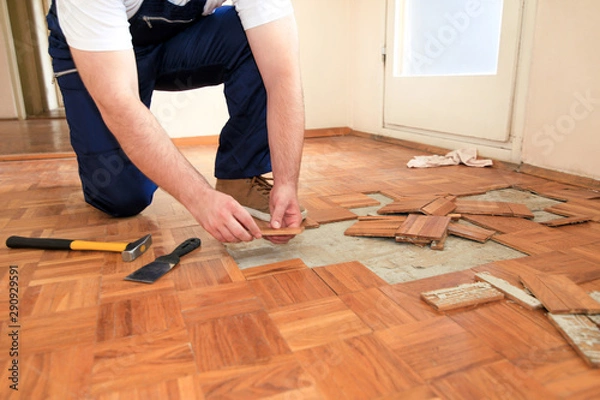 Fototapeta Construction worker and constructor is working on renovation of apartment. Handyman is removing old wooden parquet flooring using yellow hammer and scraping tool. Builder dismantles parquet floor.