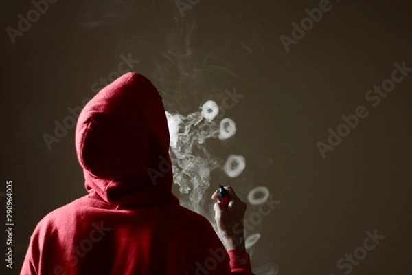 Fototapeta Young male in red hoodie vaping smoking, blows multiple smoke rings while holding a vape in hand, isolated rear view