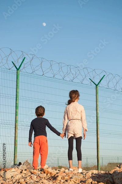 Obraz poor refugees migrant kids on state border with high fence with barbed razor wire and moon in sky holding hands with hope for care