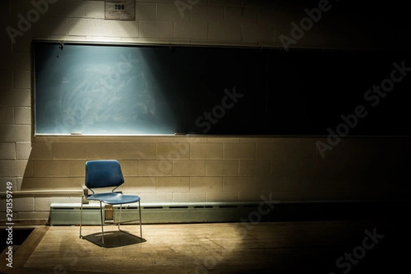 Fototapeta An Empty School Chair in a Dark, Shadowy Classroom - in Front of a Chalkboard with a Single Beam of Light Overhead