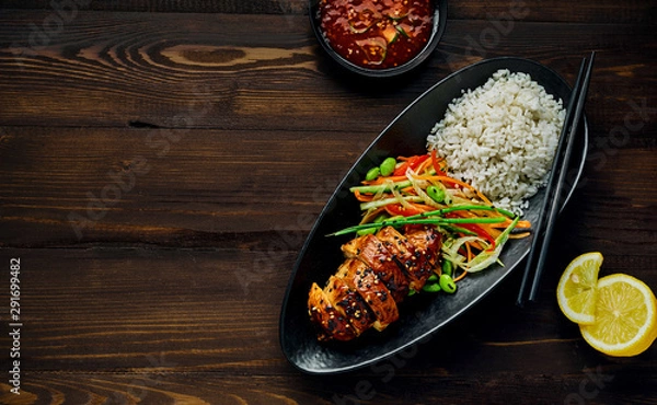 Obraz Teriyaki chicken with cucumber, ginger, cabbage and carrot salad, edamame soy beans and basmati rice in a black pottery dish with sweet chili sauce and lemon. Top view, directly above.