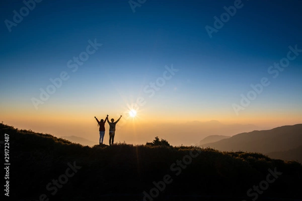 Fototapeta silhouette of a couple with arms raised up showing their happiness and successful with background of mountains and golden sunshine .